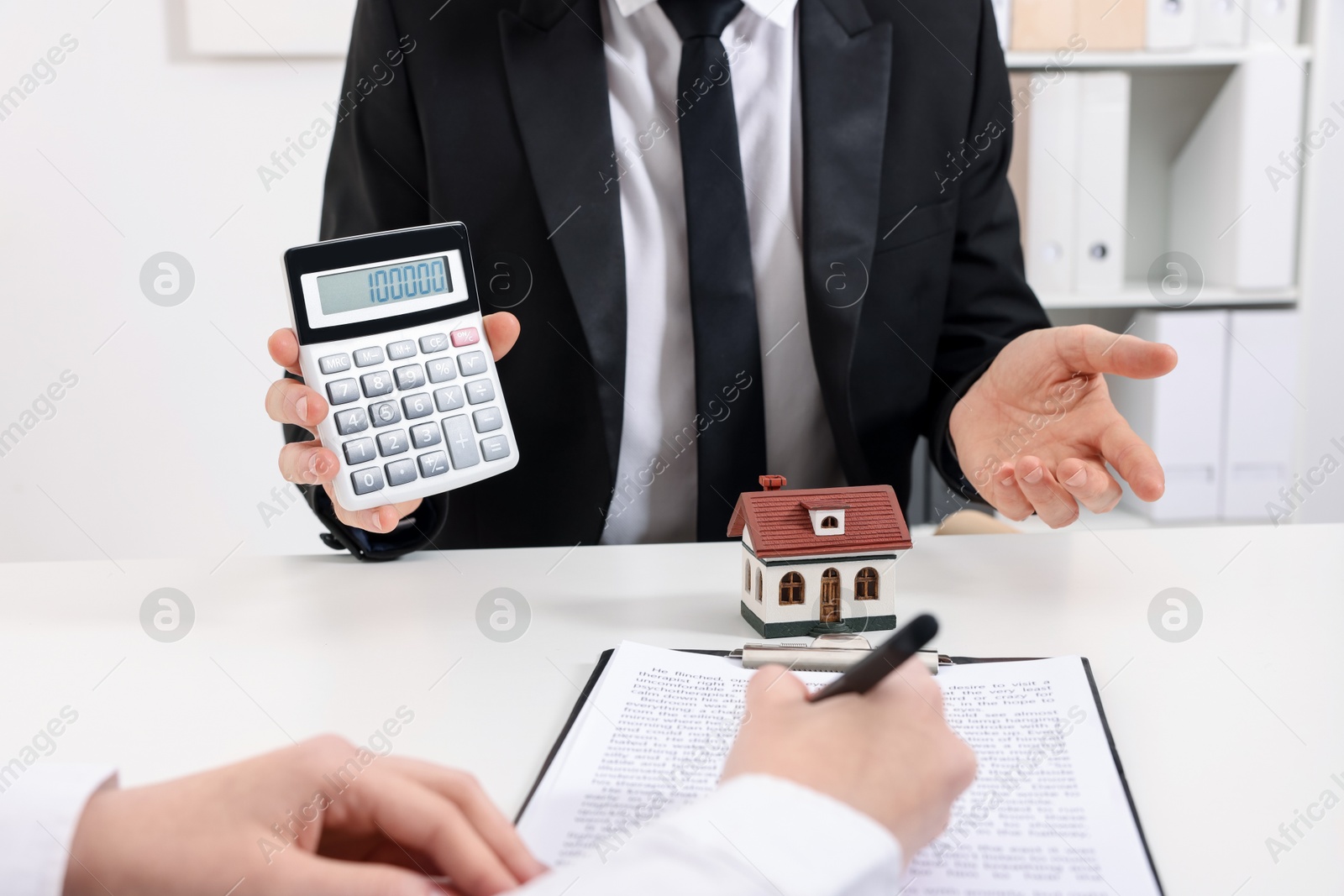 Photo of Real estate agent showing calculator with price for house while client signing document at white table in office, closeup. Home appraisal