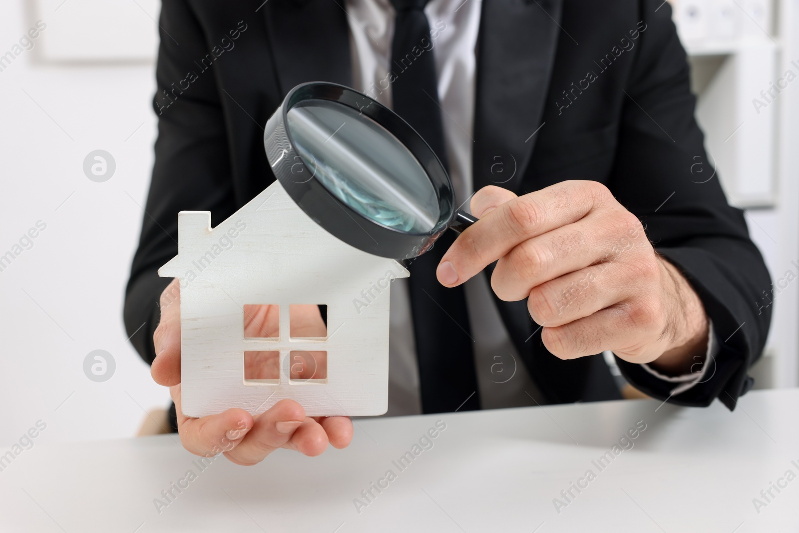 Photo of Real estate agent with house figure and magnifying glass at white table in office, closeup. Home appraisal