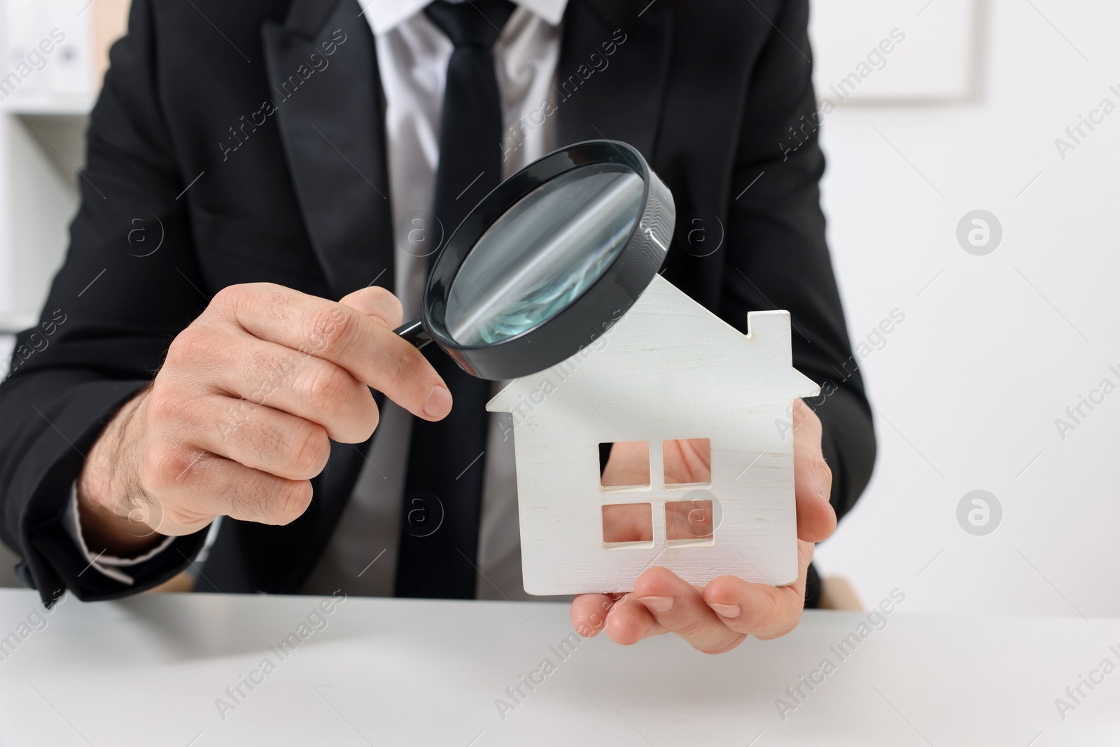 Photo of Real estate agent with house figure and magnifying glass at white table in office, closeup. Home appraisal