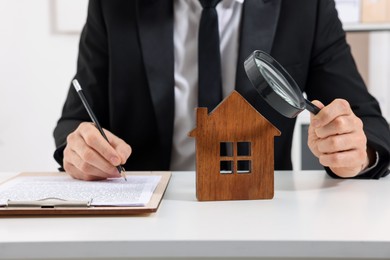 Photo of Real estate agent with house figure and magnifying glass signing document at white table in office, closeup. Home appraisal