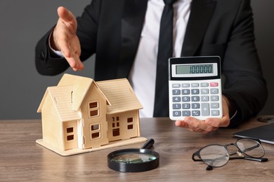 Photo of Real estate agent showing calculator with price for house at wooden table against grey background, closeup. Home appraisal