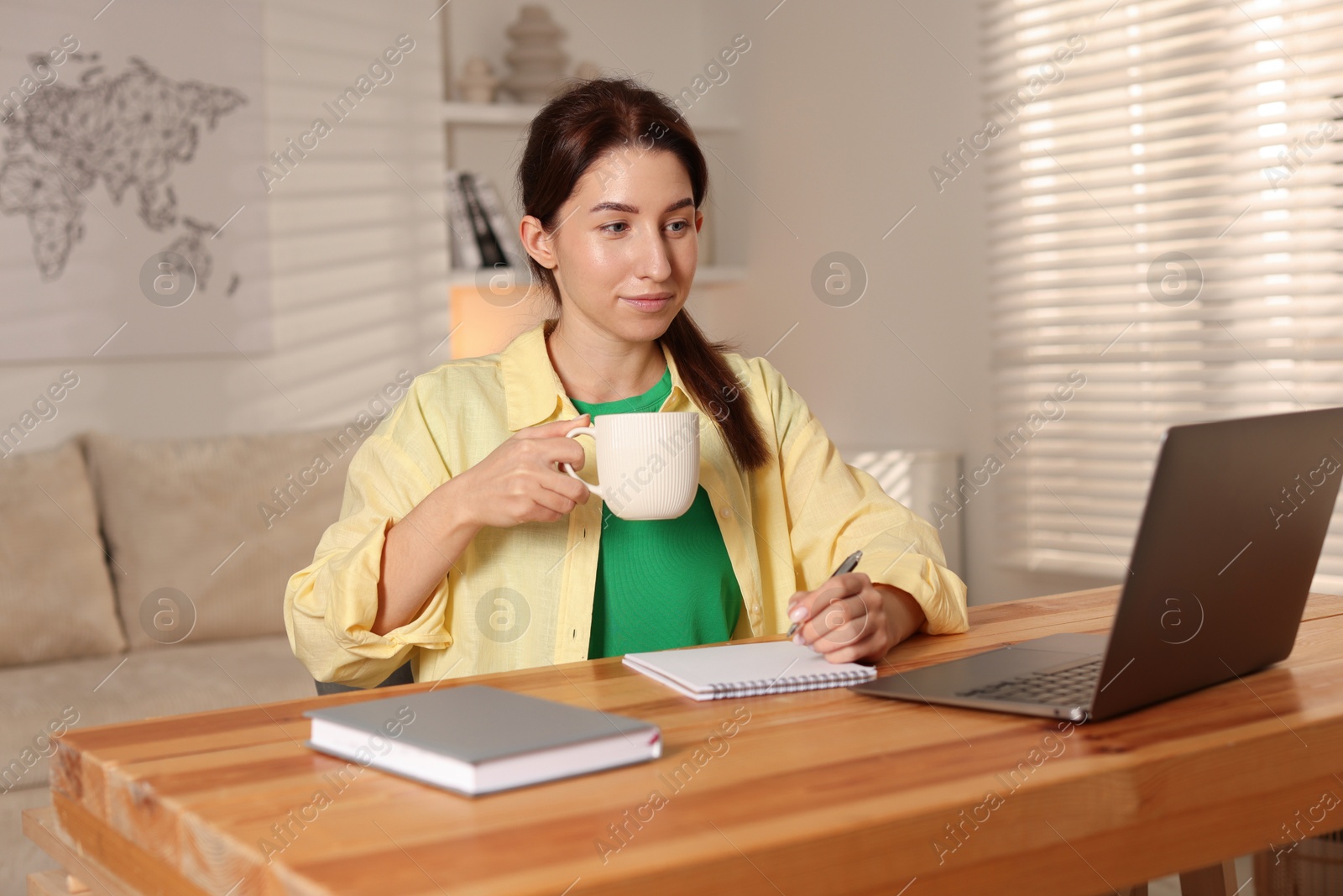 Photo of Young woman with cup of coffee working on laptop at desk in home office