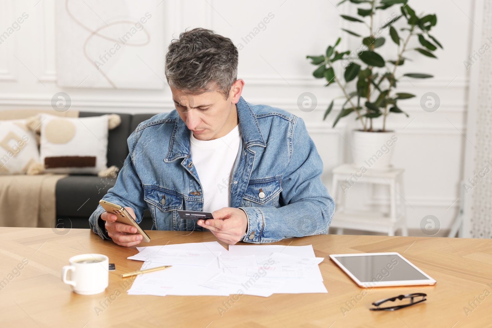 Photo of Man paying bills with credit card via smartphone at wooden table indoors