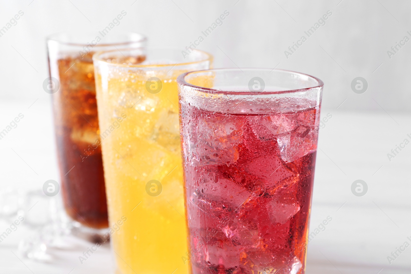 Photo of Soda water of different flavors with ice cubes in glasses on white table, closeup