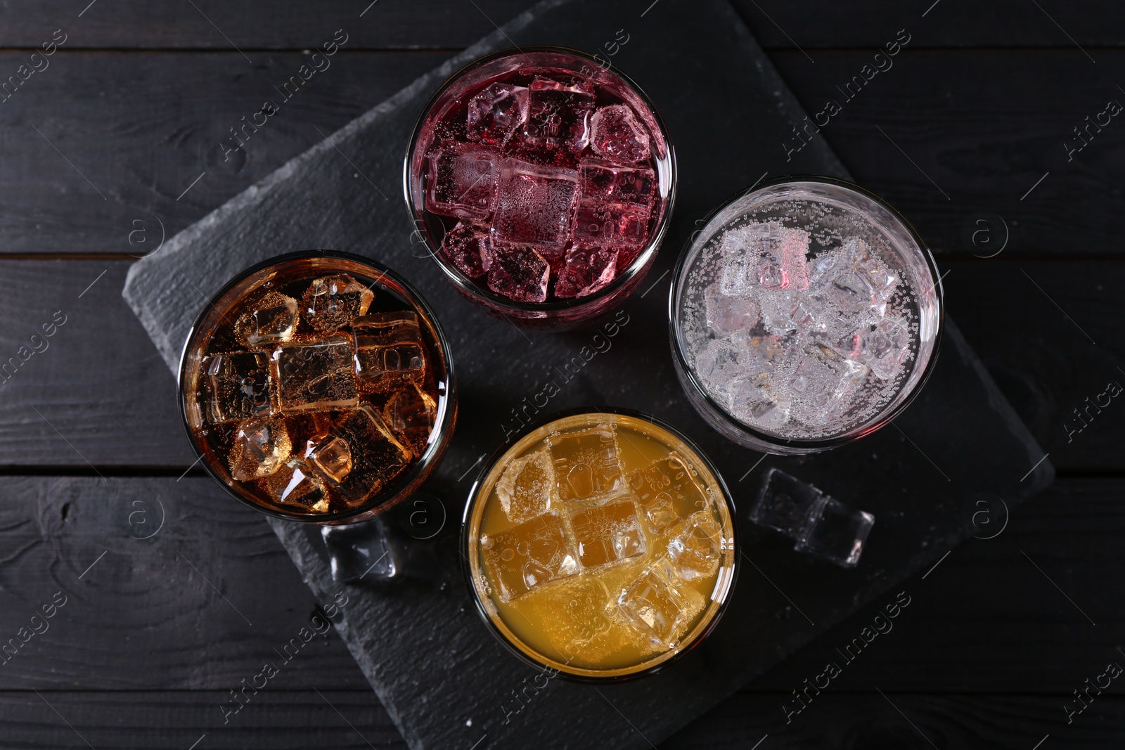 Photo of Refreshing soda water of different flavors in glasses with ice cubes on black wooden table, flat lay