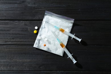 Photo of Drug addiction. Plastic bag with powder, syringes and pills on black wooden table, top view