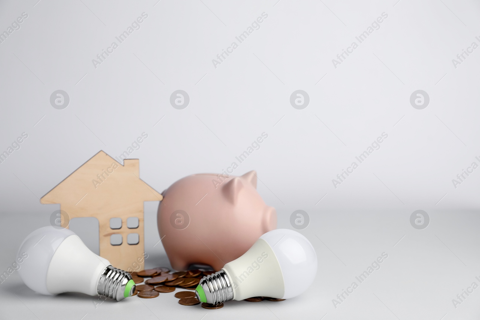 Photo of Piggy bank, coins, house model and light bulbs on grey background, space for text. Energy saving concept