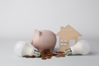 Photo of Piggy bank, coins, house model and light bulbs on grey background. Energy saving concept