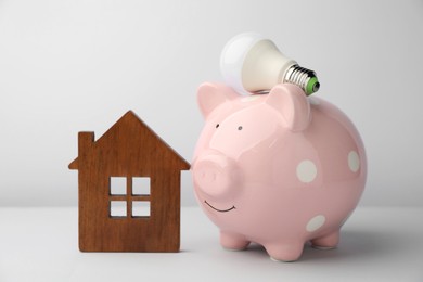 Photo of Piggy bank, house model and light bulb on grey background. Energy saving concept