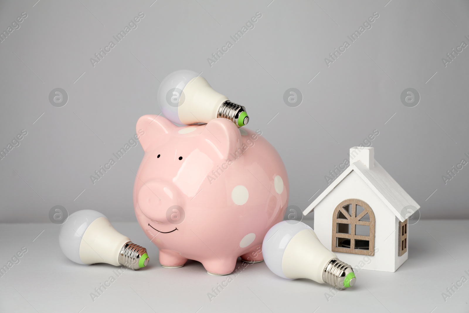 Photo of Piggy bank, house figure and light bulbs on grey background. Energy saving concept