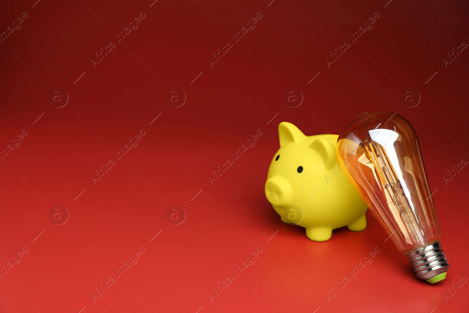 Photo of Piggy bank and light bulb on red background, space for text. Energy saving concept