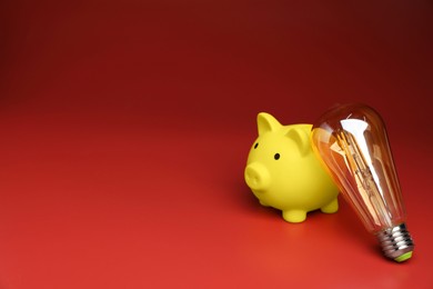 Photo of Piggy bank and light bulb on red background, space for text. Energy saving concept