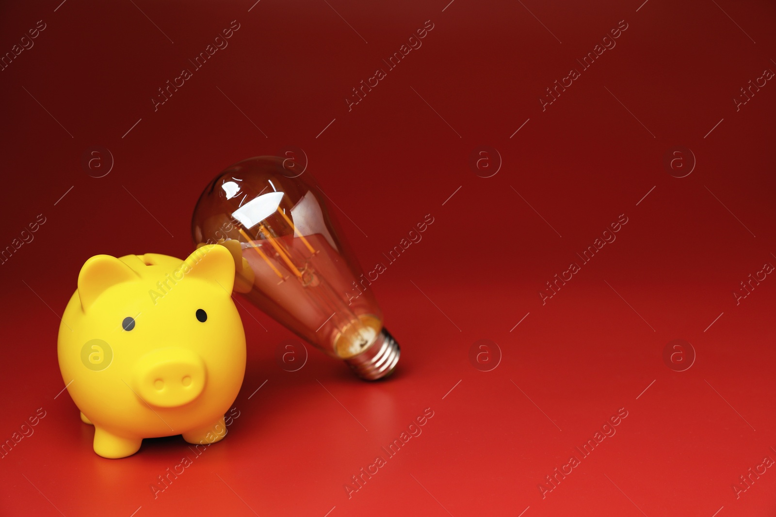 Photo of Piggy bank and light bulb on red background, space for text. Energy saving concept