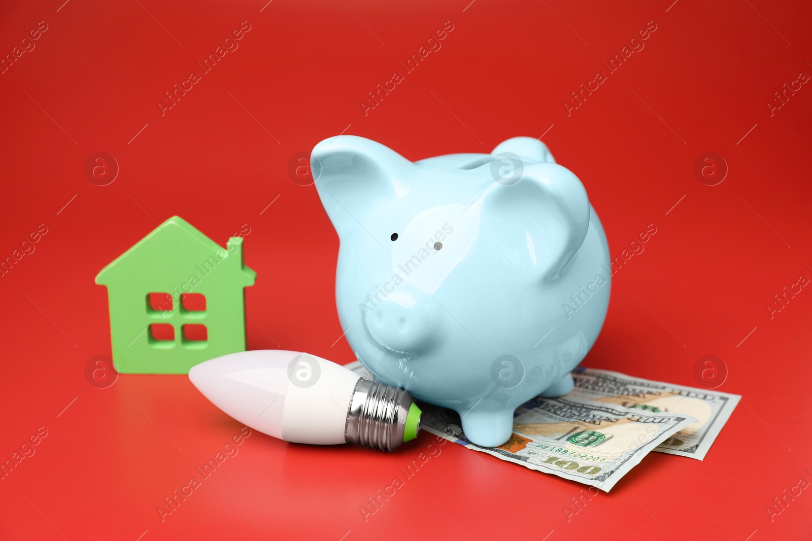 Photo of Piggy bank, dollar banknotes, house model and light bulb on red background. Energy saving concept