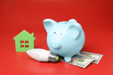 Photo of Piggy bank, dollar banknotes, house model and light bulb on red background. Energy saving concept