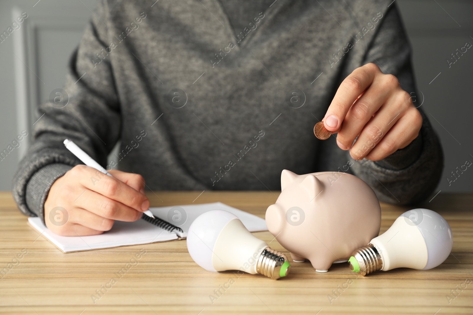 Photo of Man putting coin into piggy bank while taking notes at wooden table, closeup. Energy saving concept