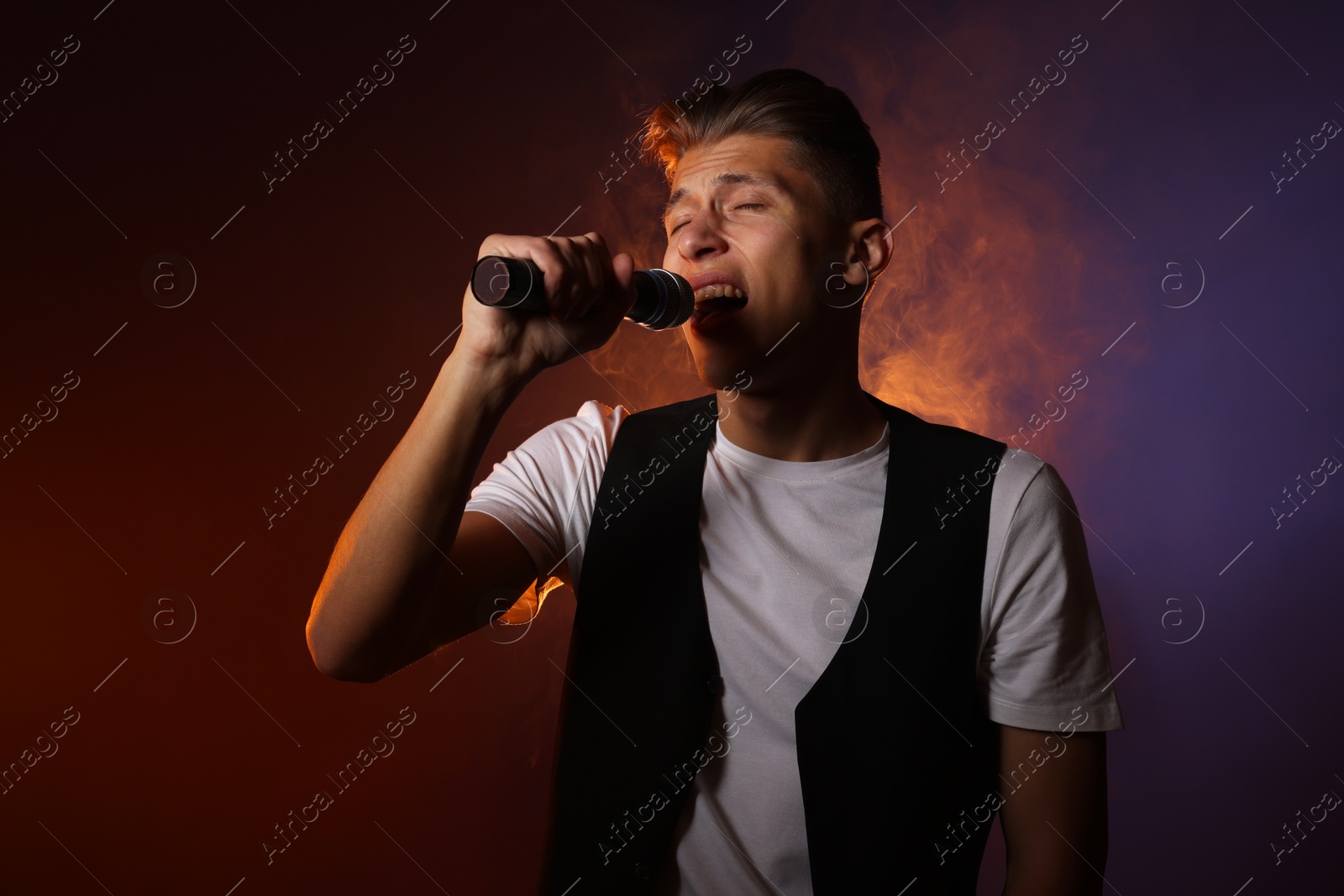 Photo of Talented young man singing on dark background with color light and smoke