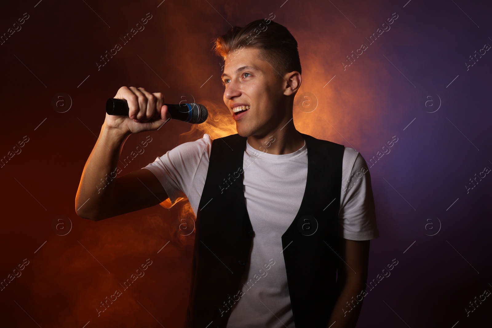 Photo of Talented young man singing on dark background with color light and smoke