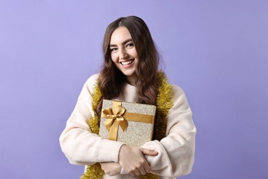 Photo of Happy young woman with tinsel and gift box on purple background