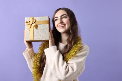 Photo of Happy young woman with tinsel and gift box on purple background