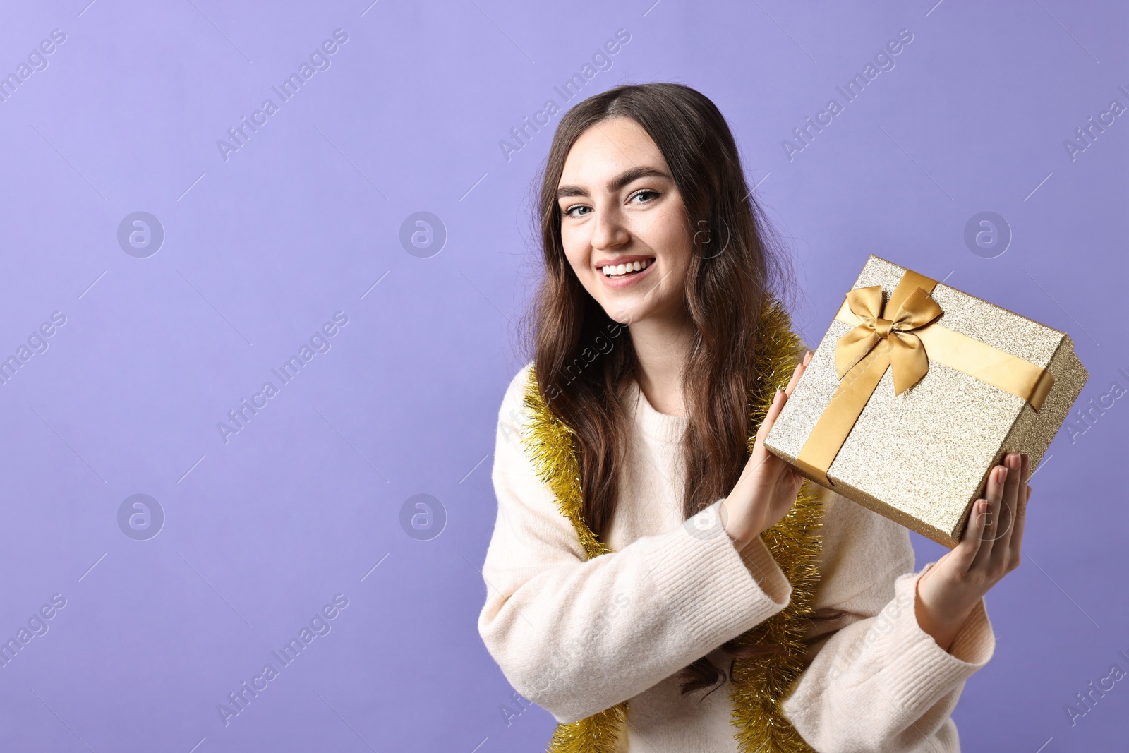 Photo of Happy young woman with tinsel and gift box on purple background. Space for text