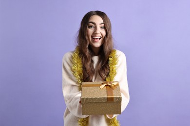 Photo of Happy young woman with tinsel and gift box on purple background