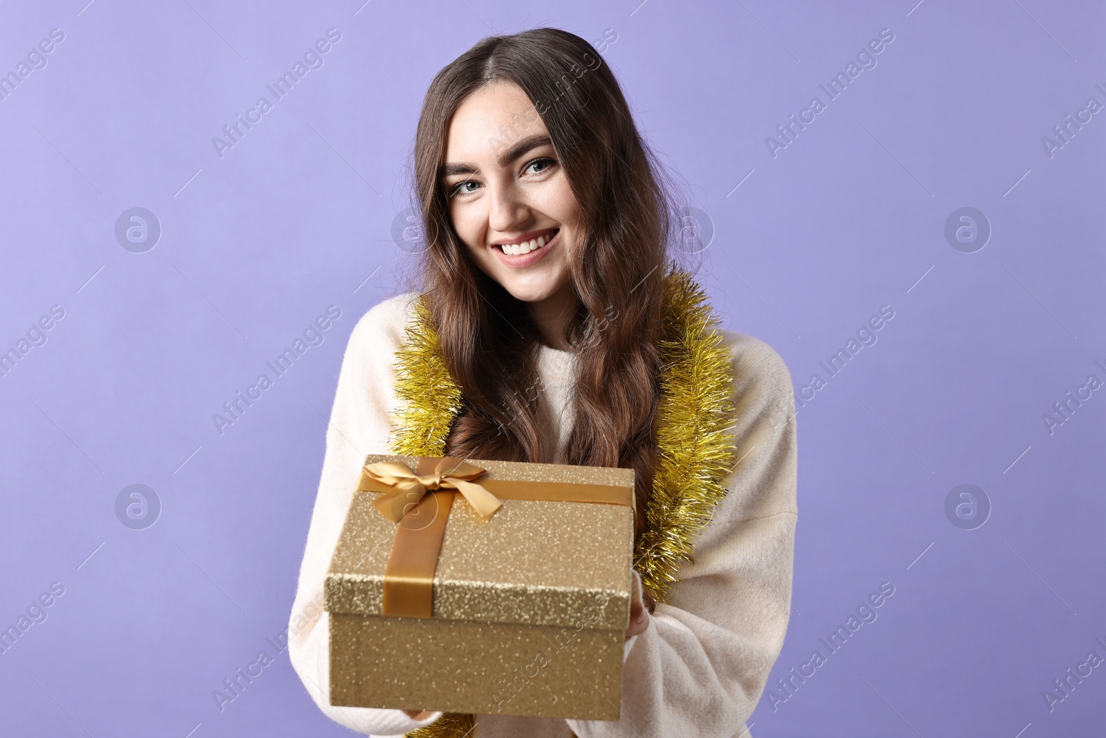 Photo of Happy young woman with tinsel and gift box on purple background