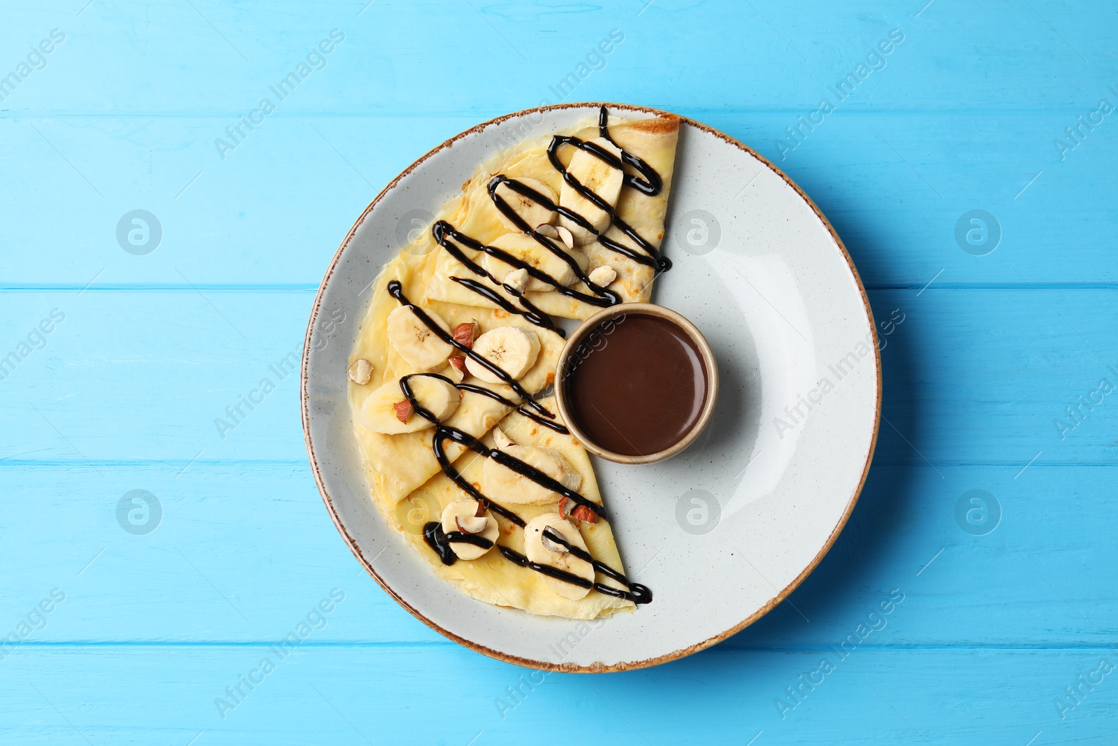 Photo of Delicious crepes with banana, nuts and chocolate sauce on light blue wooden table, top view