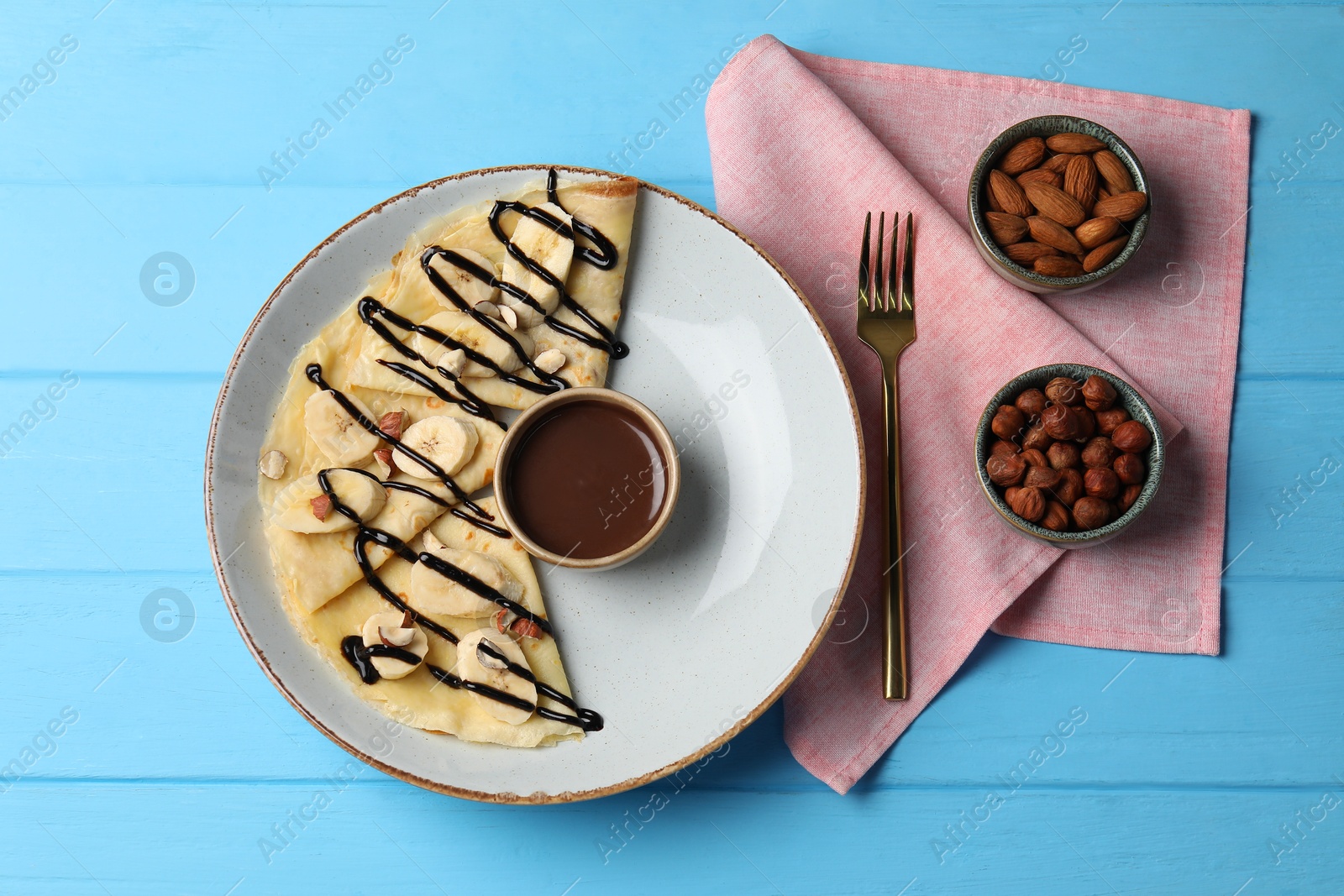 Photo of Delicious crepes with banana, nuts and chocolate sauce on light blue wooden table, flat lay