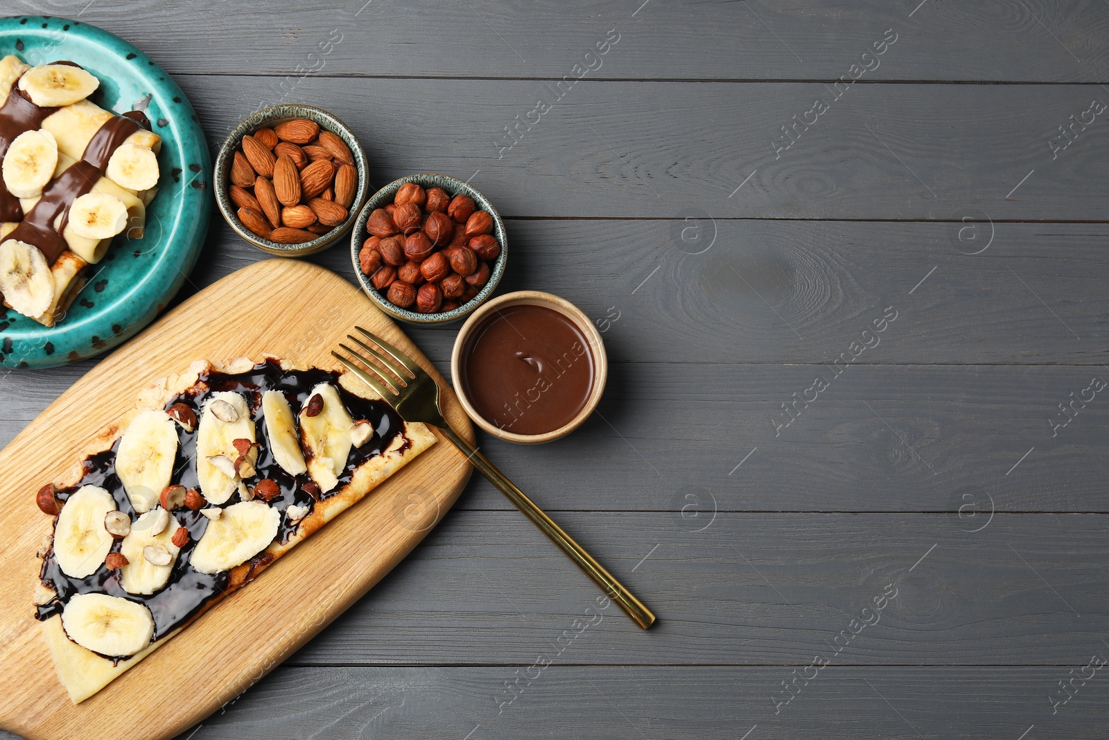 Photo of Delicious crepes with banana, nuts and chocolate sauce on grey wooden table, flat lay. Space for text