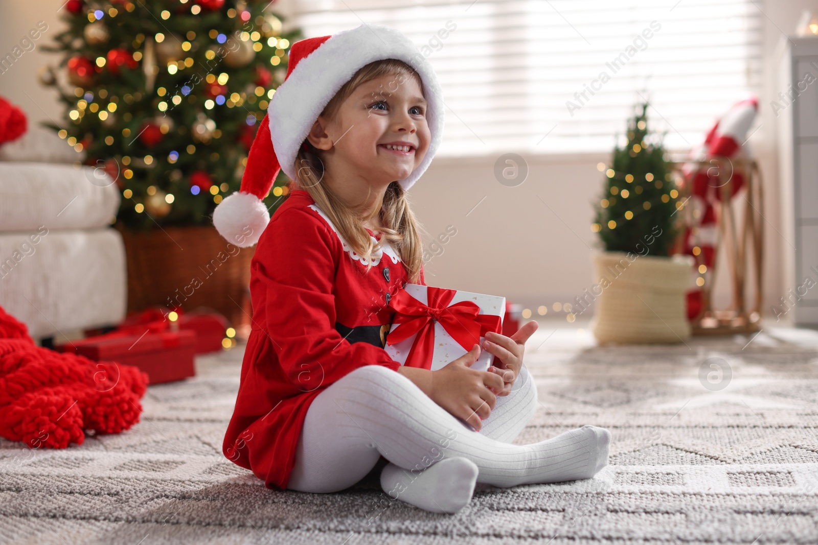 Photo of Little girl in Santa hat with gift box sitting on floor at home