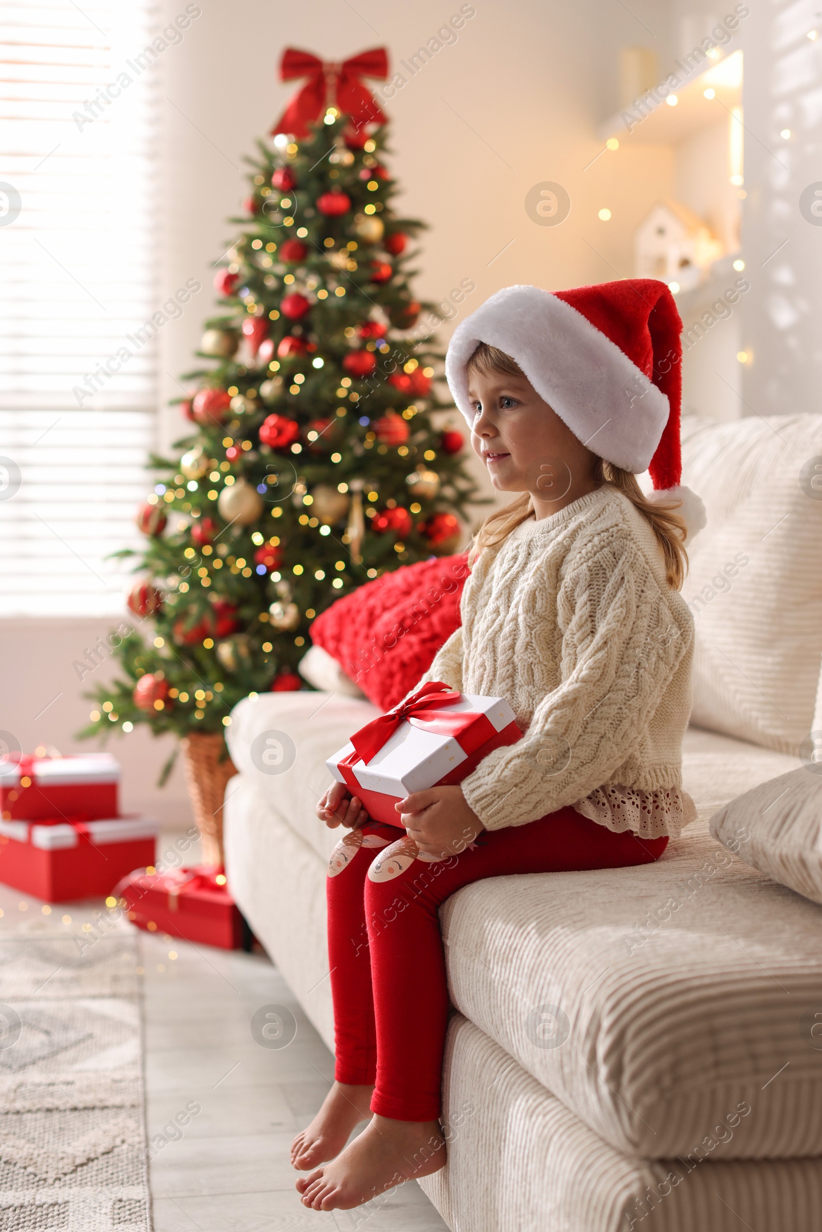 Photo of Little girl in Santa hat sitting on sofa at home