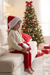 Photo of Little girl in Santa hat sitting on sofa at home