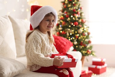 Photo of Little girl in Santa hat sitting on sofa at home