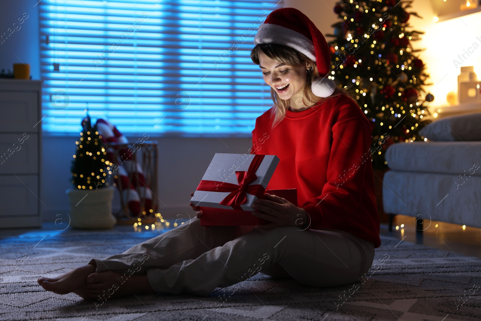 Photo of Happy woman in Santa hat with Christmas gift on floor at home