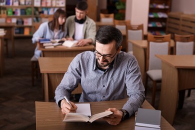 Handsome man reading book and taking notes at desk in public library