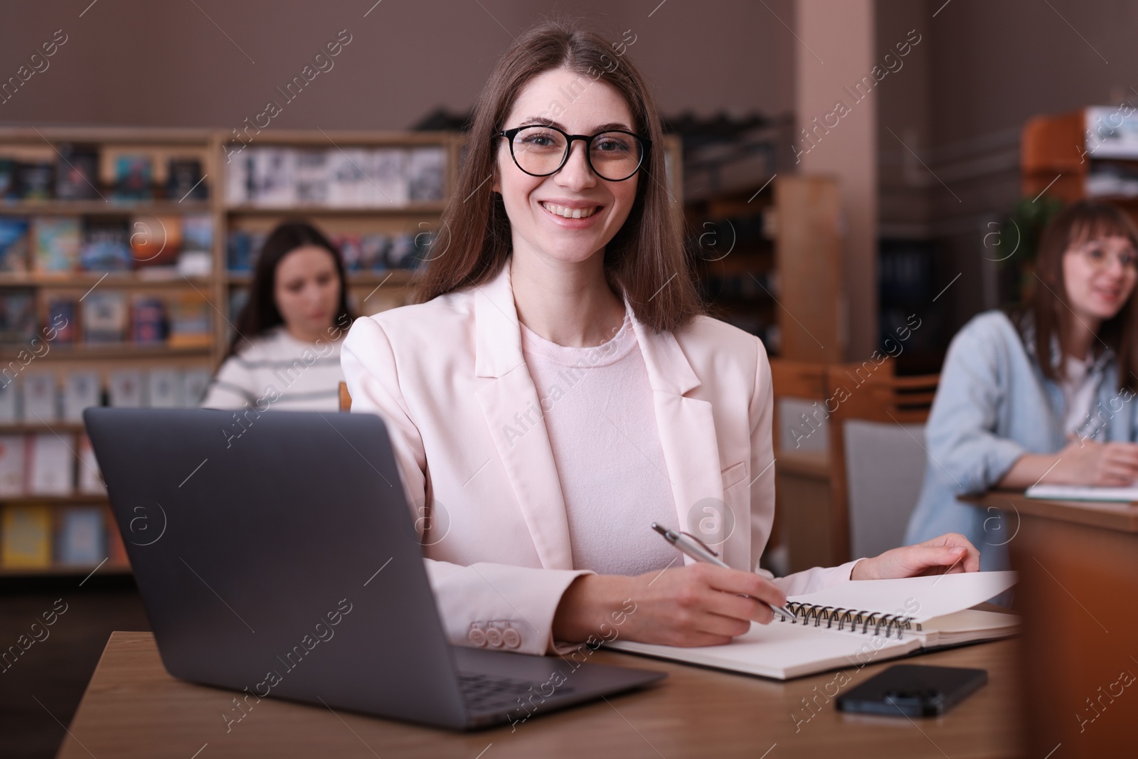 Photo of Portrait of smiling woman with laptop taking notes at desk in public library