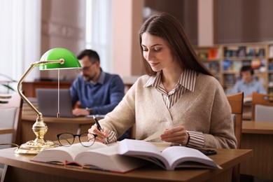 Photo of Beautiful woman reading book and taking notes at desk in public library