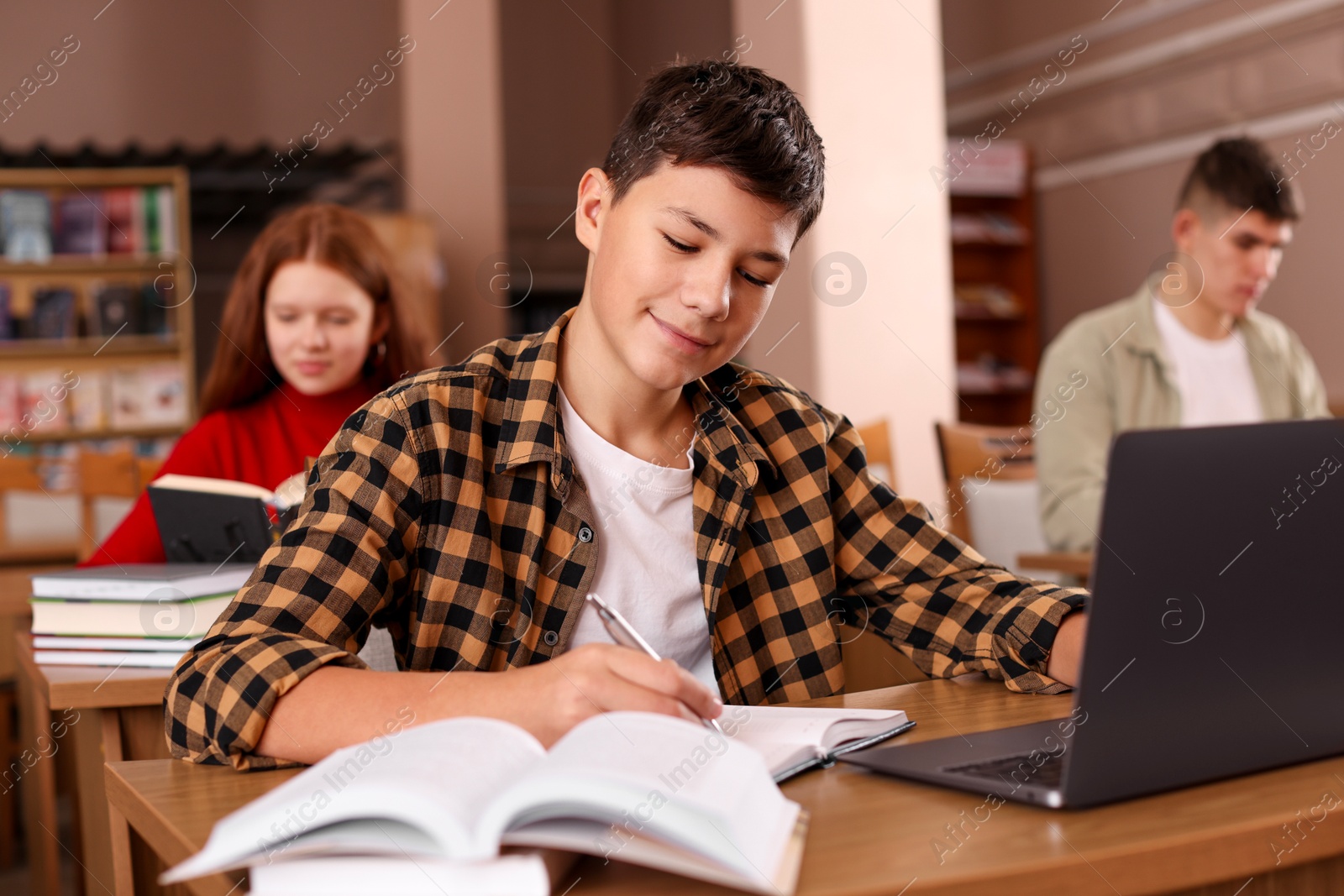 Photo of Boy taking notes while using laptop at desk in public library