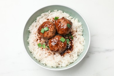 Photo of Delicious rice with meatballs, sauce and green onions in bowl on white marble table, top view