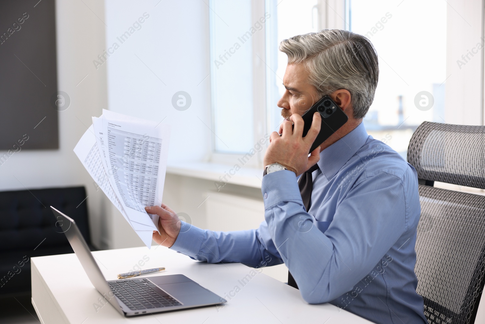 Photo of Banker with documents talking on smartphone at desk in office