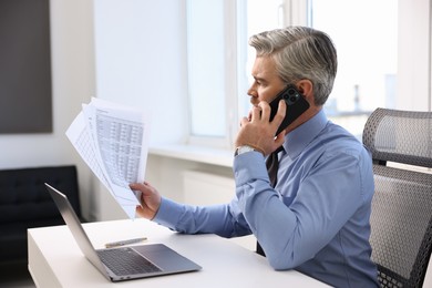 Photo of Banker with documents talking on smartphone at desk in office