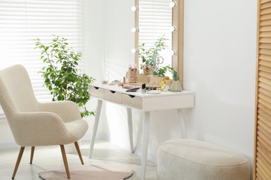 Photo of Makeup room. Dressing table with mirror, different beauty products, jewelry, armchair and houseplant indoors