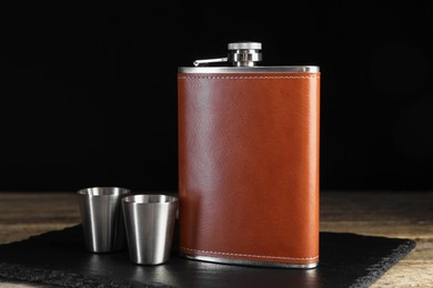 Photo of Hip flask and cups on wooden table against black background