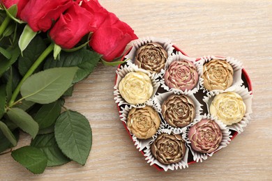 Photo of Flower shaped chocolate bonbons in box and red roses on wooden table, flat lay