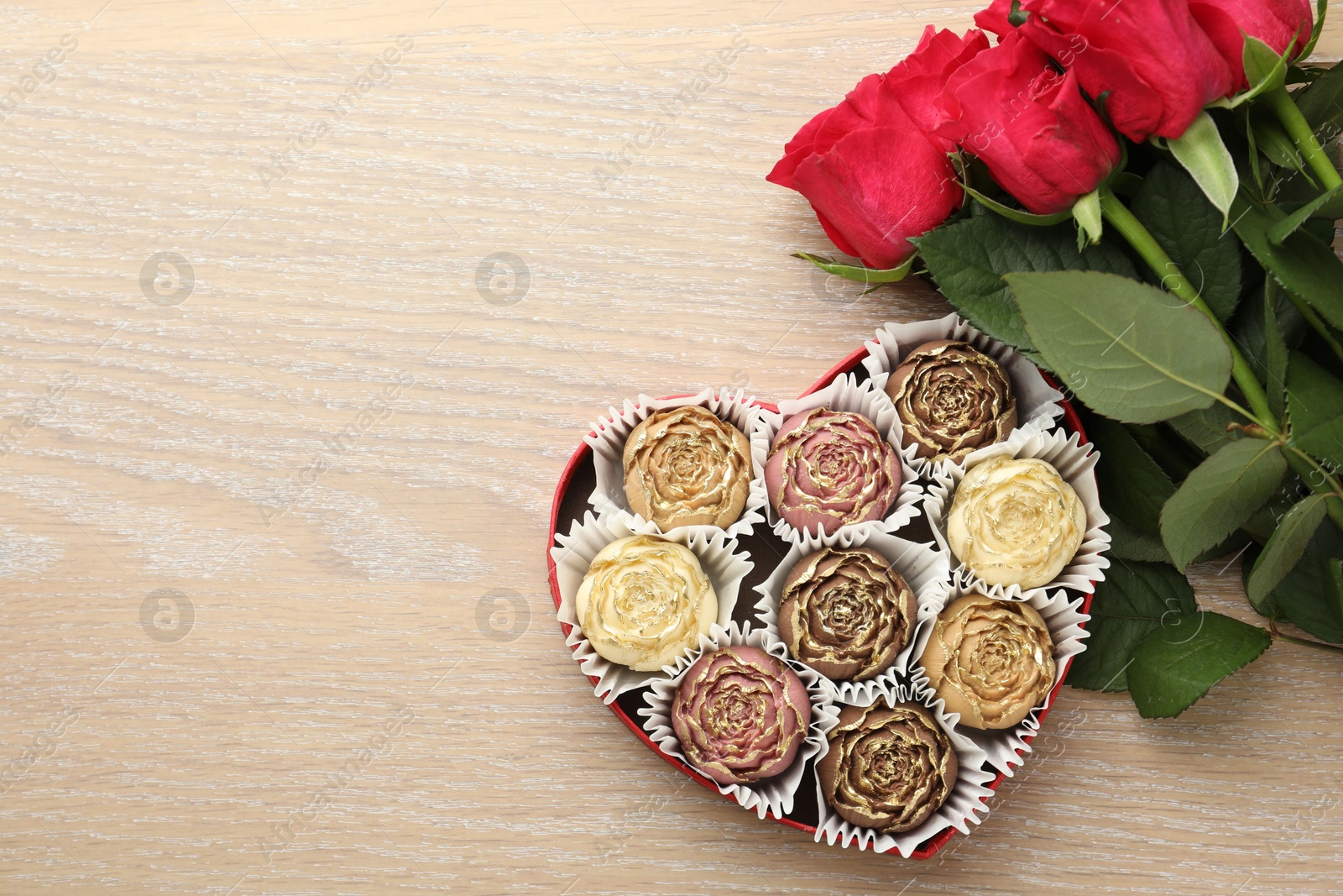 Photo of Flower shaped chocolate bonbons in box and red roses on wooden table, flat lay. Space for text