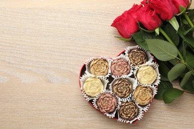 Flower shaped chocolate bonbons in box and red roses on wooden table, flat lay. Space for text