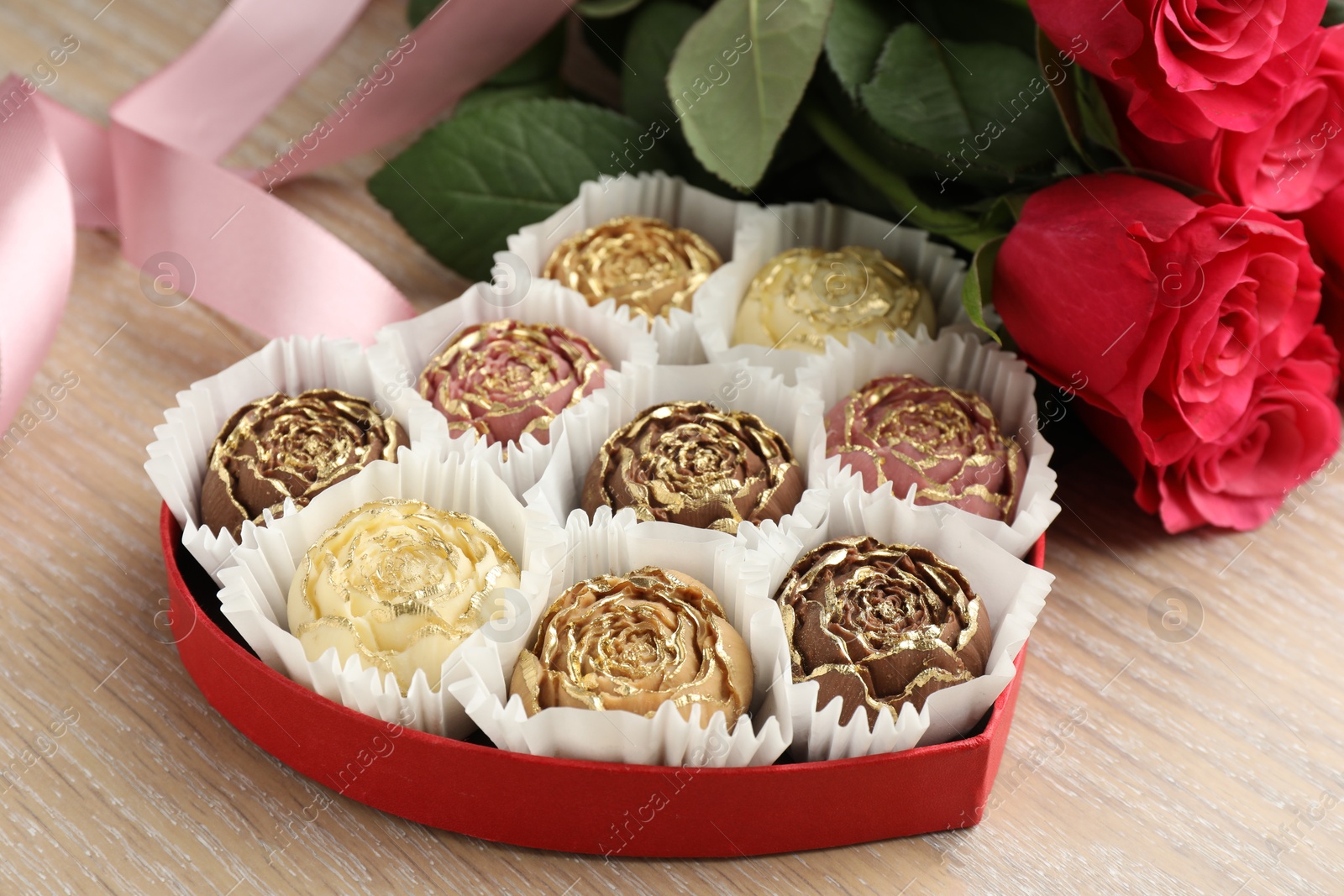 Photo of Flower shaped chocolate bonbons in box, red roses and pink ribbon on wooden table, closeup