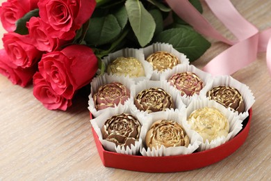 Flower shaped chocolate bonbons in box, red roses and pink ribbon on wooden table, closeup