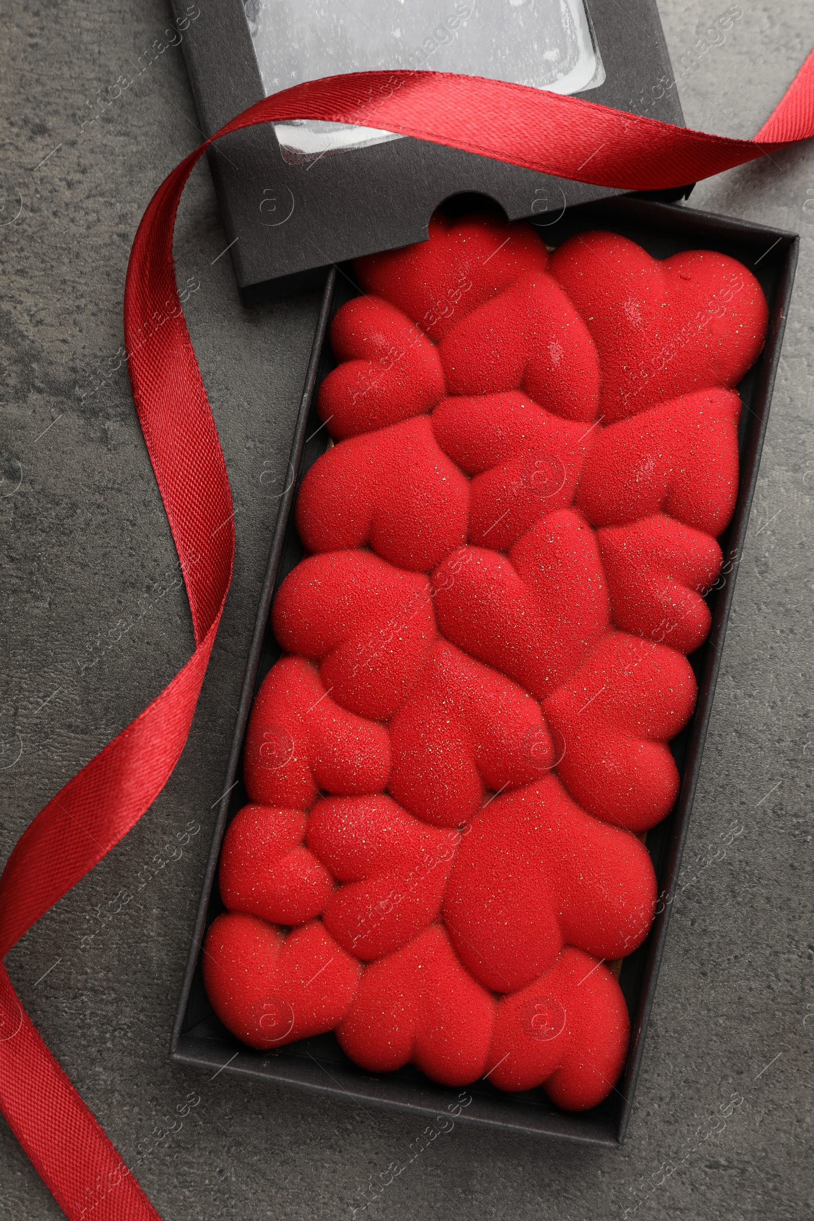 Photo of Chocolate bar decorated with hearts in box and red ribbon on grey table, flat lay